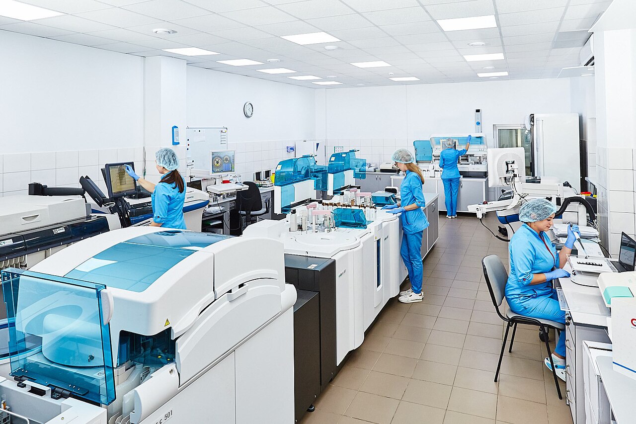 The image shows a group of scientists working in a laboratory. The scientists are wearing lab coats and gloves. They are working on a variety of different tasks, including looking 
